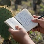 Manos de un estudiante con un cuaderno de campo junto a un cactus de tuna