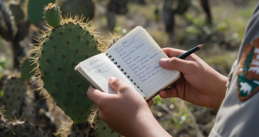 Manos de un estudiante con un cuaderno de campo junto a un cactus de tuna
